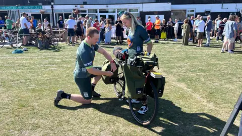 Ciaran Booth kneels beside a loaded touring bike while Lucy Pickersgill stands next to him on a grassy rugby ground. There are people milling behind them, but they area the focus of the shot.