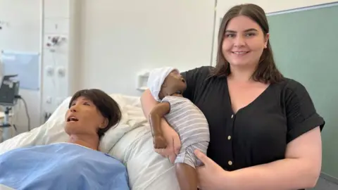 A lady wearing a black top is smiling holding a plastic baby wearing a grey striped babygrow. She is stood next to a hospital bed, with a dummy patient who symbolises a mother who has just given birth.