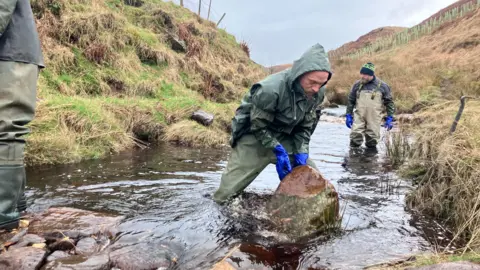 A man wearing green waterproof gear in a stream handling a large piece of stone.