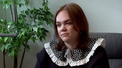 Emily Dove sits on a chair. She is wearing a navy blue top with a polka dot-patterned collar. She has shoulder-length brown hair and a pierced nose.