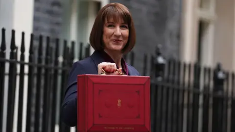 PA Chancellor Rachel Reeves holds up a red suitcase on Budget Day as she stands on Downing Street. She is slightly smiling as she looks to her right side.