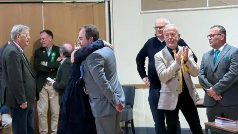 Louise Parker/BBC a man is hugging a womans he is wearing a suit and she is wearing a dress. Other candidates are clapping 