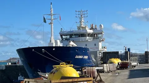 A large blue-and-white ship named Sarnia Cherie is moored at a concrete harbour dock under a bright blue sky. Yellow industrial buoys and equipment sit in the foreground, while the vessel’s bridge, antennas, and masts rise above the deck.