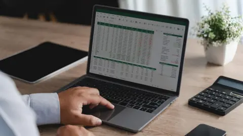 Getty Images Person reviewing data and graphs in spreadsheet documents on a laptop with a calculator.