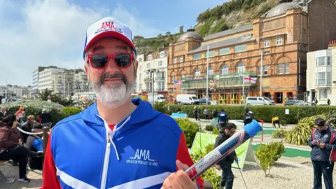 ROBIN GREY/BBC A man dressed in matching red, white and blue zip-up jumper and cap, holding a golf club. He is stood at a crazy gold course in Hastings and is also wearing sunglesses.