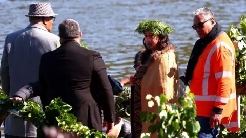 Getty Images Maori Queen Kuini Nga wai hono i te po stands next to a river wearing a headress and a cloak, with three men standing alongside her.