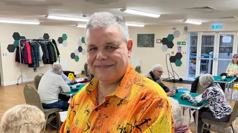 A man with short cropped grey hair, wearing a bright orange and yellow shirt , stood in a room with people playing bridge behind him 