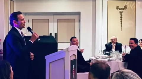 Terry Pattinson Mark stands at a podium holding a microphone, addressing an audience seated at tables. The tables are covered with white tablecloths and set with glassware and plates.