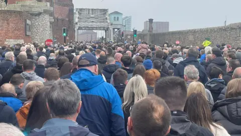 Thousands of Everton fans walk along Regent Road, on the right is a large stone dock wall and on the left is a brick wall with a sign for the Titanic Hotel