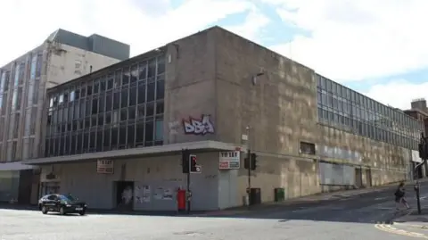 A derelict building with 'To Let' signs on either side of the corners. There's graffiti on the walls and boarding up windows around the bottom of the building. 