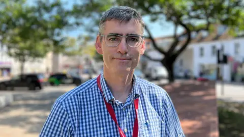 Jersey deputy Montfort Tadier smiles while stood in a public square in St Helier. He has glasses on and is wearing a blue chequed shirt. He has a red lanyard round his neck and has short greying hair. It is a sunny day.