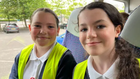 Image of two schoolgirls seated inside a bus. They are wearing high- viz vests over their school uniforms
