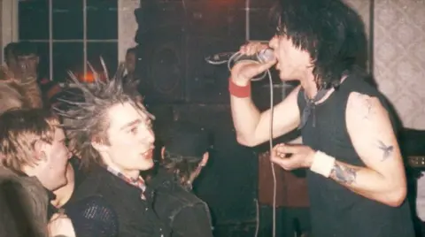 Historic England/Home of Metal A man with shoulder-length black hair sings into a microphone, with speakers in the background and people in the crowd watching him, including a man with spiky hair