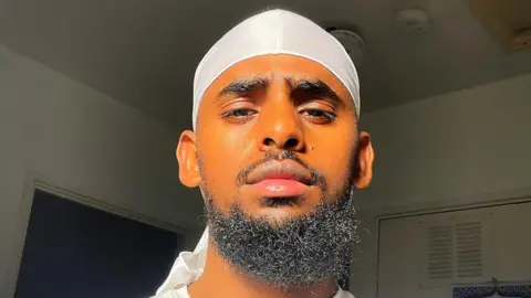 Bahar Telila Man with curly beard, looks early twenties. Poses for photo in sunlight in front of a window. He wears a white head covering in the selfie