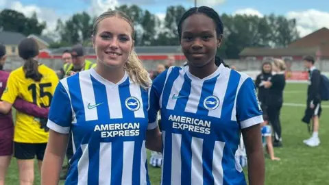 Isla Hubbard and Nazaraei Forbes with one arm around each other as they pose for a photo in their white and blue striped jerseys. Isla has her blond hair tied back and Nazaraei has her black hair tied back.