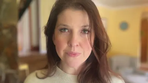 Katy wears a cream coloured jumper with straight dark brown long hair