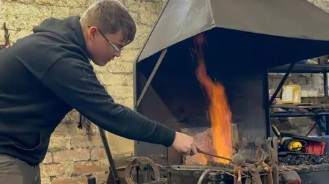 BBC Felix leaning over the forge, stoking a fire. He's wearing a black hoodie, grey trousers and protective googles. In the foreground you can see an anvil and a hammer, with various pieces of smithing equipment hanging off the forge. Behind the forge is an exposed brick wall, with hammers hanging from a hook. 