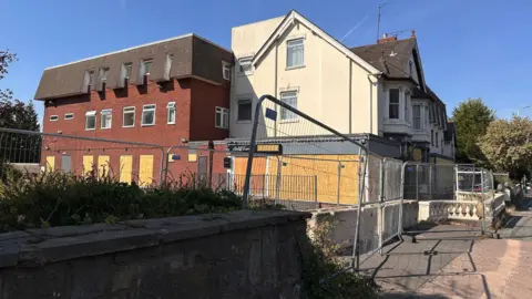 One side of the hotel site dominates the image, with some of it mainly yellow and another section mainly red on the outside. A second side of the hotel, at right angles to the most visible side, is on the far right of the photo, with silver barriers in front of the building.