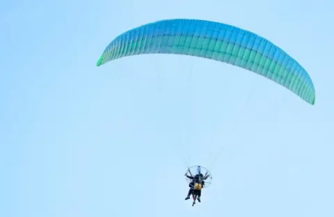 Getty Images/Ali Cobanoglu Paramotor with blue parachute and two people riding it.