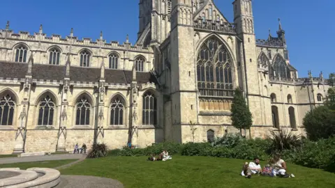 Gloucester Cathedral pictures from the side on a sunny day. People are lounging on the grass around the cathedral, which is lit up by the sun under a blue sky.