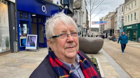 A man stood in a pedestrianised street in a commercial town centre. He is wearing glasses, a dark coat and a multi-coloured check scarf. In the background, a clock can be seen mounted above one of the buildings.