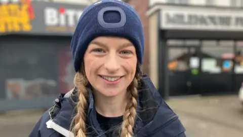 A woman, wearing a blue woollen hat and a blue coat. She has plaited hair, and is smiling at the camera.