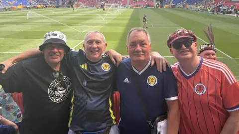 Four men with their arms around each others' shoulders inside a football stadium, with the pitch and other stands visible behind them. The two men in the middle are wearing blue Scotland tops. The man on the left is wearing a black t-shirt and a bucket hat, and the man on the right has a salmon pink Scotland top and a tartan cap and sunglasses.