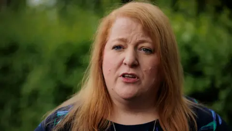 PA Media Headshot of Naomi Long speaking outside with some bushes in the background. She is wearing a navy, blue and green top.