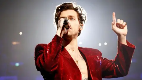 Harry Styles on stage. He is wearing an open red velvet jacket, and holding one finger in the air