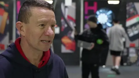 Karl Holden with short brown hair wearing a red and navy hoodie in his darts shop with darts boards and customers behind him.