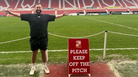 Man stood near football pitch which says Swindon behind