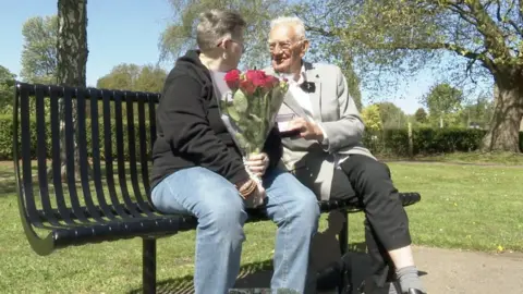 The photo shows a man and a woman sitting on a black metal bench. The woman is wearing a black hoodie and blue jeans. The man is wearing a grey blazer and black trousers. The woman is holding a bunch of red roses. They're sitting in a park. 