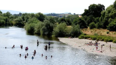 Reuters People swim in the River Wye during a heatwave in July 2022. You can see the water and swimmers in the forefront, people on a pebbly beach to the right, and greenery in the background.