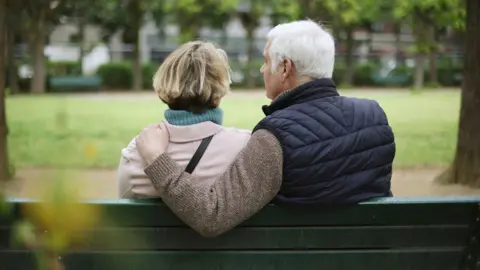 An older couple sit on a park bench. The man has his arm around the woman and there are trees and grass in the background. 