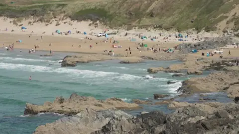 Fistral Beach is a wide sandy beach with lots of people walking and lying on it and there are people swimming in the sea alongside jagged rocks. There are many windbreaks and beach tents. It is a slightly cloudy day.