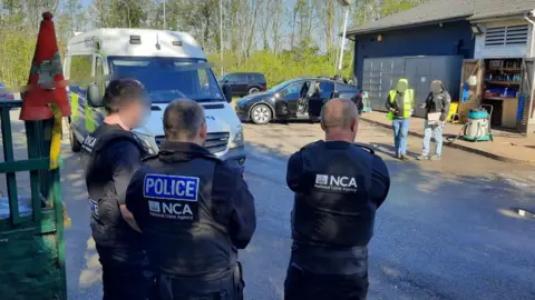 NCA officers watch as other officials and officers question staff at a industrial site