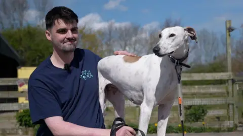 Jack and Mojo, a white lurcher style dog sit side by side. Jack has short black hair and a faint black beard.
