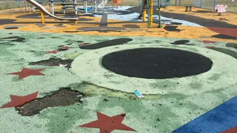 A children's play area with a green, yellow and blue rubber style floor surface. Large holes have been ripped out of the floor showing the black material beneath. 