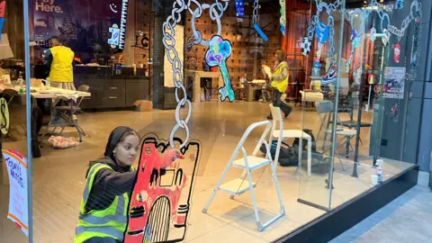 Jon Wright/BBC A girl paints a mural on a shop window, with a large red medieval gate along with chains and keys