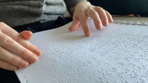 A white page of braille with Evie's right hand resting on the page on the left, and her fingers of her right hand running over the bumps on the page, to the right.