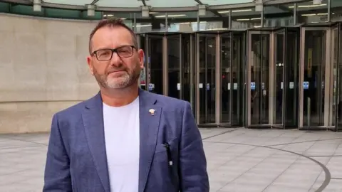 David Clayton is wearing glasses. He has short dark hair and a short beard and is wearing a blue suit jacket over a white T-shirt.