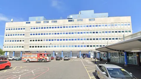 The long, grey concrete building is six storeys tall and has blue blocks on the top. There is a fire engine parked out front in the car park which is also dotted with cars. A smaller building on the right of the frame has a: "Welcome to University Hospital of North Tees" sign.