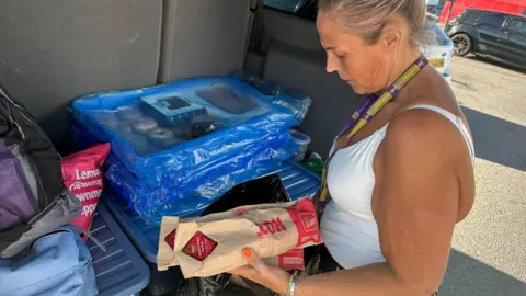 A woman in a white vest packing sandwiches in brown bags into a shopping bag in the boot of a van