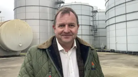 Lord John Fuller at the Brineflow site in Great Yarmouth, Norfolk, looking into the camera. He has short dark hair and is wearing a dark green jacket over a white shirt. Behind him are big metal silos.
