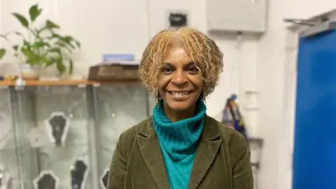Pictured Norma Banton smiling inside her jewellery studio. She has a cheek-length blonde loose curl afro. She is wearing a turquoise roll up jumper and a green corduroy blazer jacket.