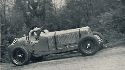 Getty Images A man is driving a vintage car in a black and white photograph of the race. The car is going up a hill and he is leaning to one side as he navigates a bend.