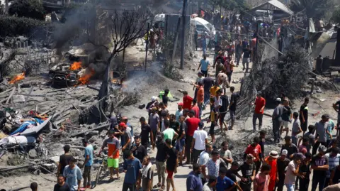 Reuters People look at the aftermath of an Israeli airstrike in Gaza. A burning car is visible to the left of the image.