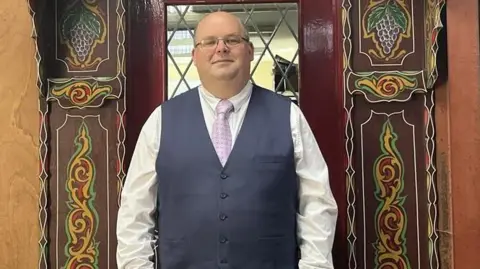 Julia Gregory/BBC The director of the museum wears a waistcoat, tie and glasses and stands in front of a brightly decorated doorway