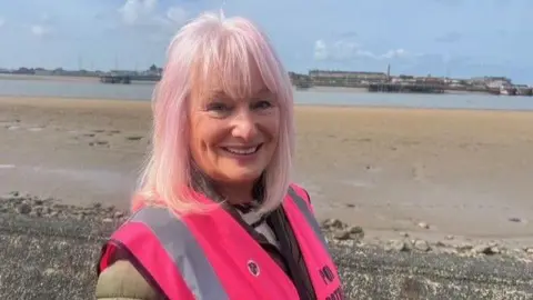 Poo Patrol organiser, Sandra Pope, wears a hi-vis pink jacket and has pink hair. She is smiling in front of a beach.