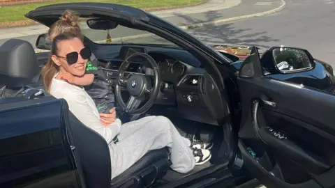 Hannah Taylor in BMW with child on lap. The door is open and she is wearing sunglasses. 